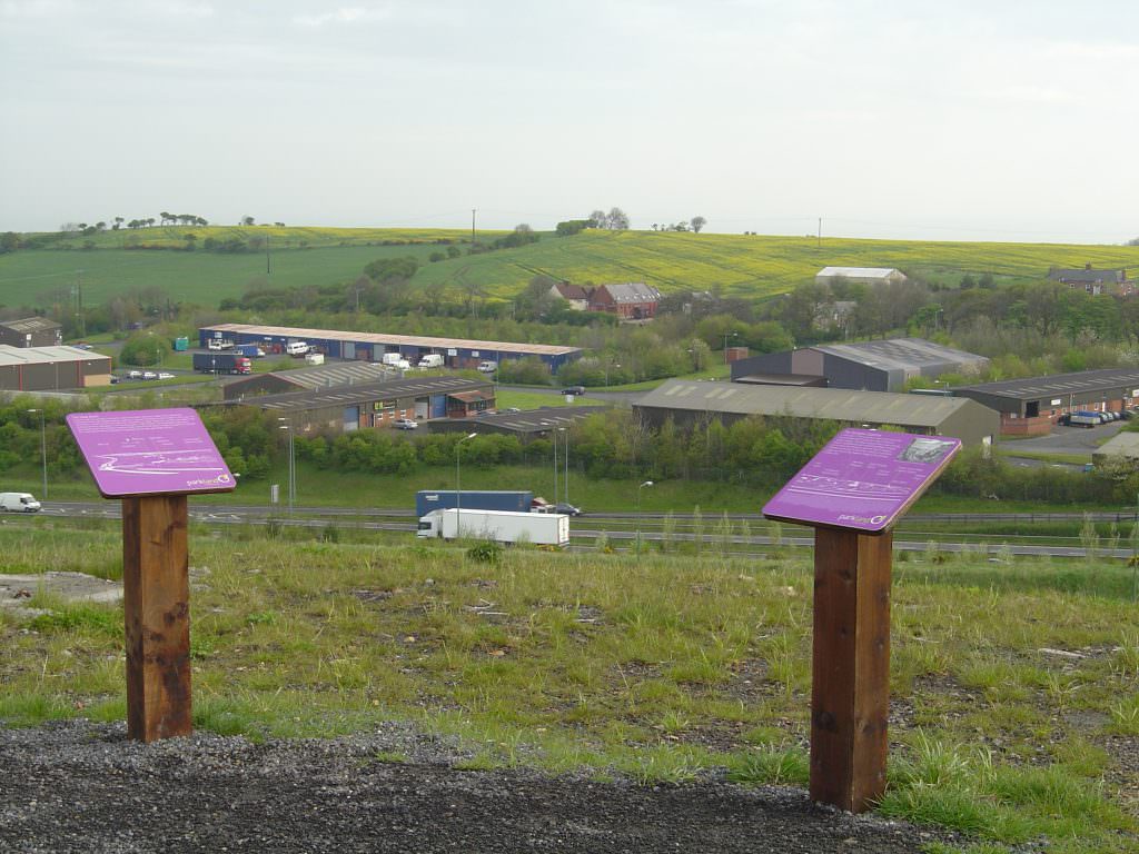 A pair of small information panels each on a single timber lectern frame. The boards are above a motorway in a new country park